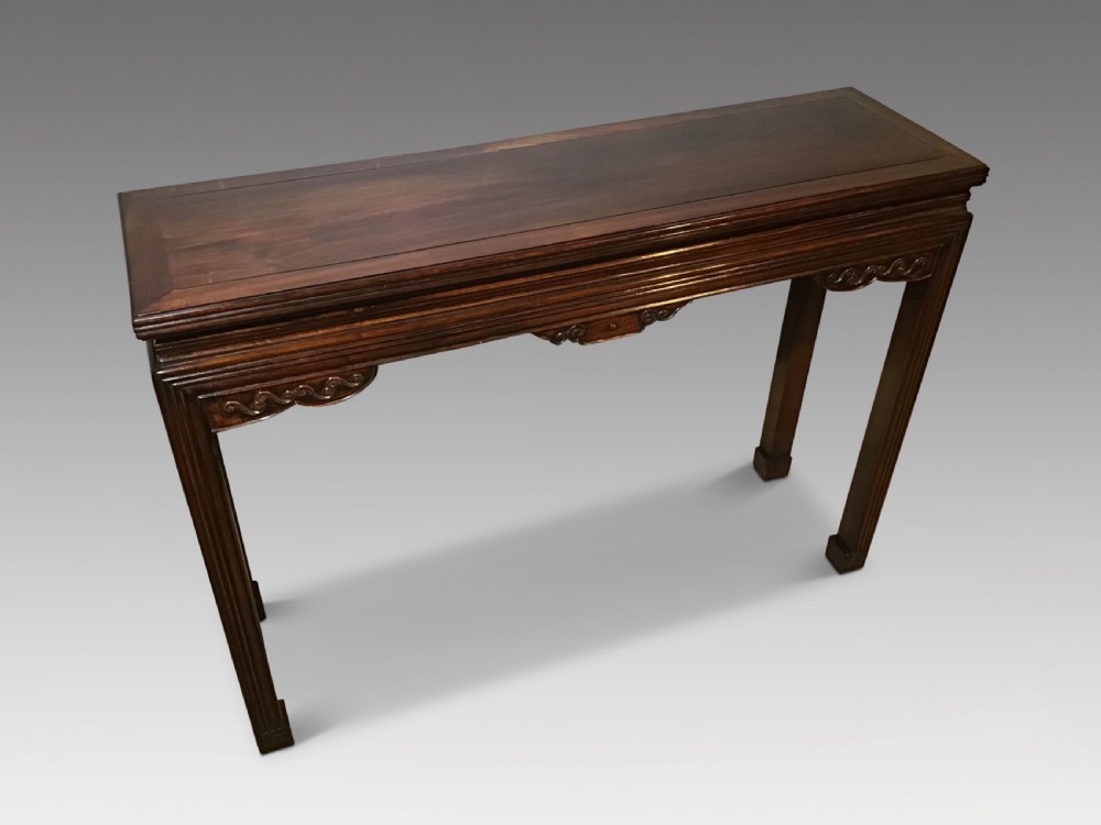 A narrow dark brown timber Chinese altar table featuring carved corner spandrels, reeded square legs, and a rectangular panelled top.