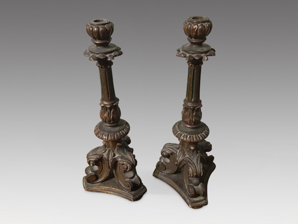 A pair of antique carved wood candlesticks featuring ornate scrolled tripod bases, fluted stems, and urn sconces with an aged dark patinated gilt finish.