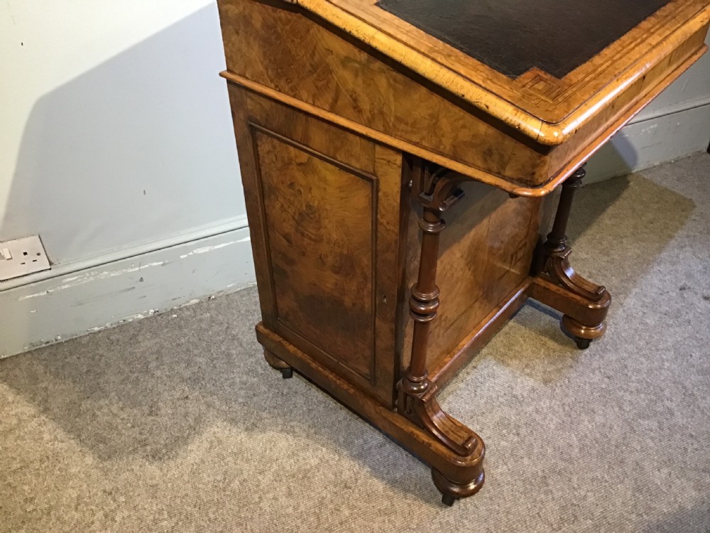 A Victorian burr walnut Davenport desk featuring a sloped leather writing surface, inlaid wood corner detailing, turned front supports, and a top stationery compartment. View V