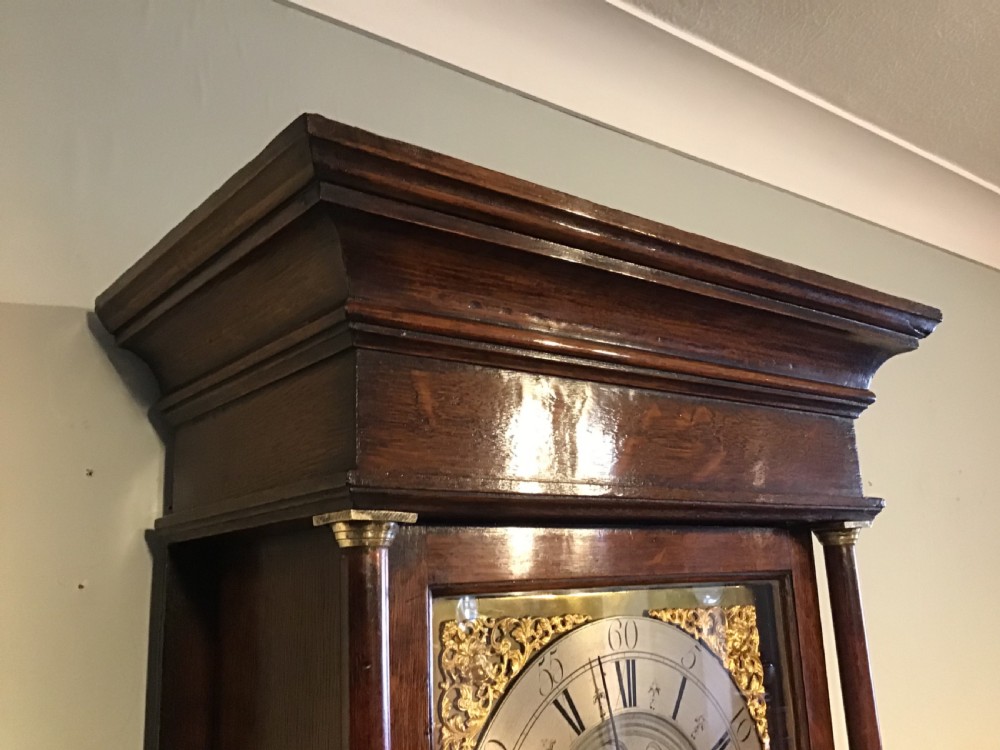 A Georgian oak eight-day longcase clock featuring an engraved brass dial, fluted hood columns, and a tall timber trunk resting on bracket feet. View VI