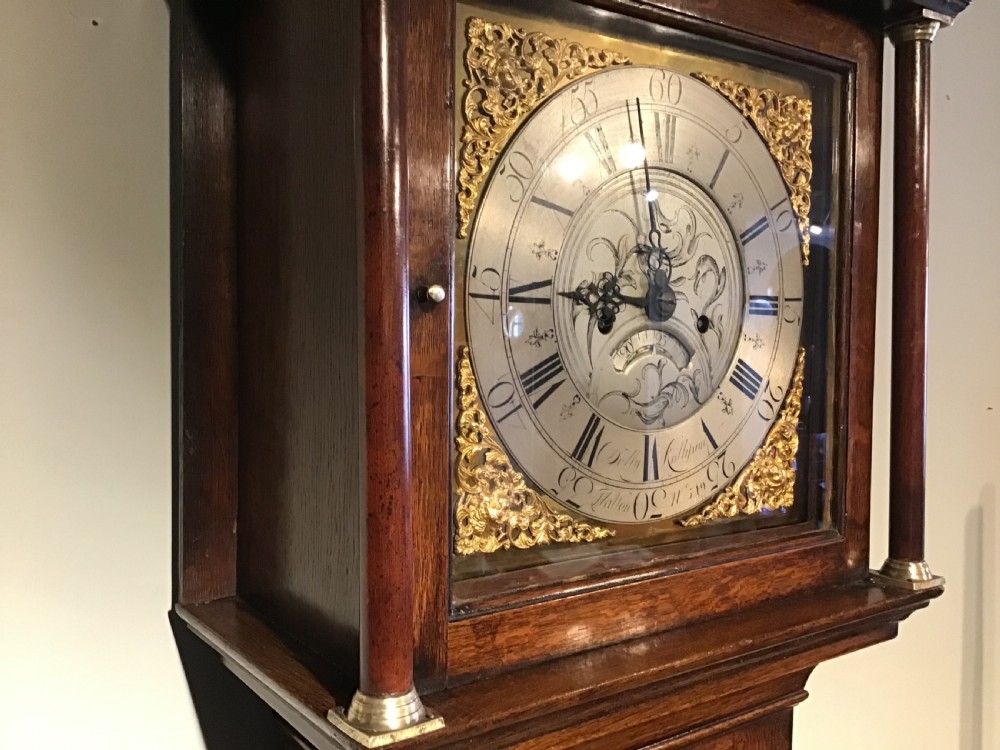 A Georgian oak eight-day longcase clock featuring an engraved brass dial, fluted hood columns, and a tall timber trunk resting on bracket feet. View VII