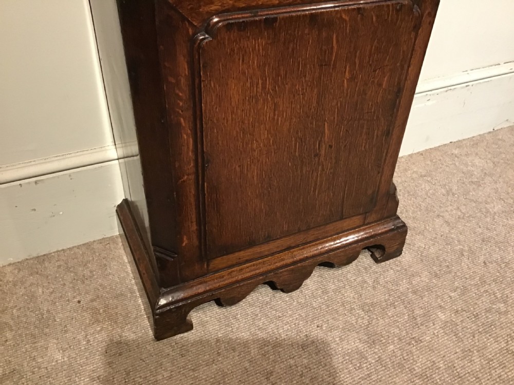 A Georgian oak eight-day longcase clock featuring an engraved brass dial, fluted hood columns, and a tall timber trunk resting on bracket feet. View X