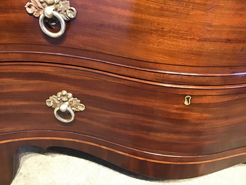 A George III style flame mahogany serpentine chest of drawers featuring four graduated drawers with brass ring handles and splayed feet. View VIII