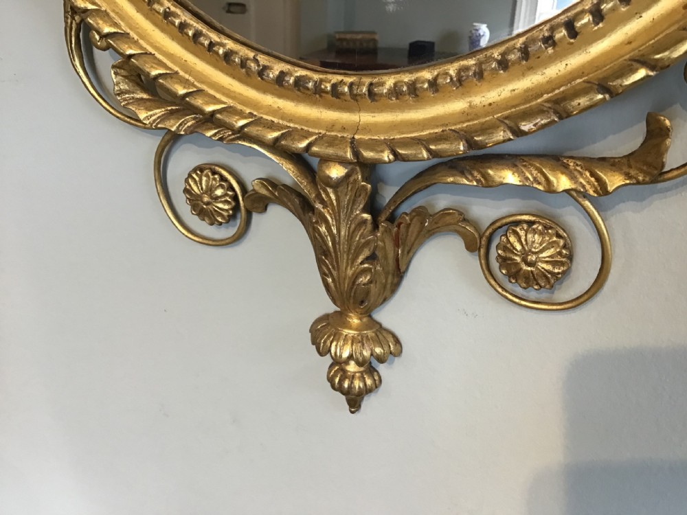 An oval wall mirror with a gilded frame topped by a classical urn finial and draped bellflower garlands along the sides. View II