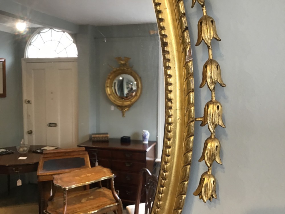An oval wall mirror with a gilded frame topped by a classical urn finial and draped bellflower garlands along the sides. View III