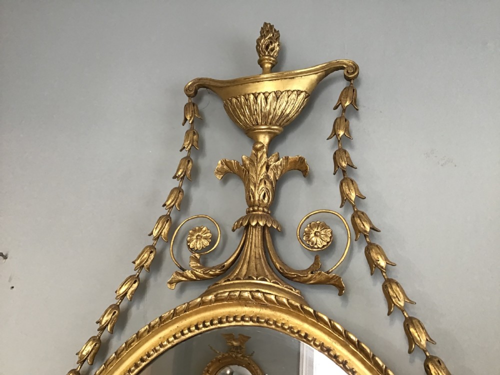 An oval wall mirror with a gilded frame topped by a classical urn finial and draped bellflower garlands along the sides. View IV