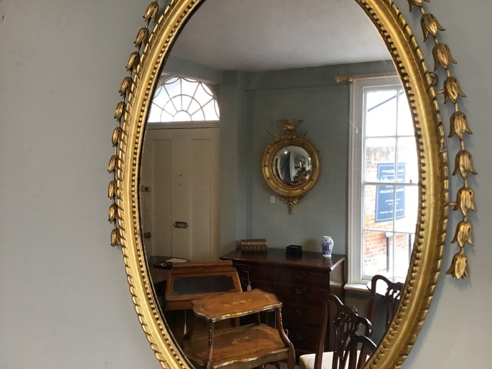 An oval wall mirror with a gilded frame topped by a classical urn finial and draped bellflower garlands along the sides. View V
