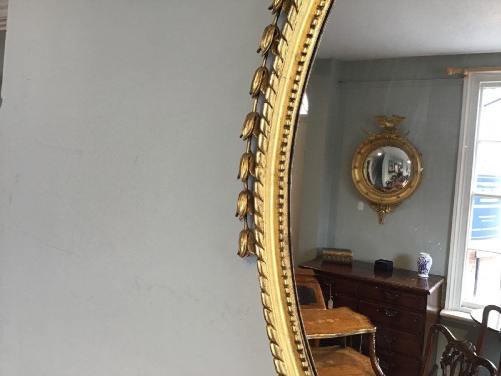 An oval wall mirror with a gilded frame topped by a classical urn finial and draped bellflower garlands along the sides. View VI