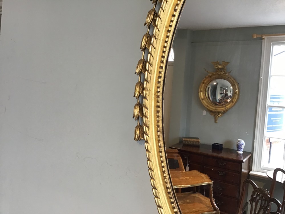 An oval wall mirror with a gilded frame topped by a classical urn finial and draped bellflower garlands along the sides. View VII