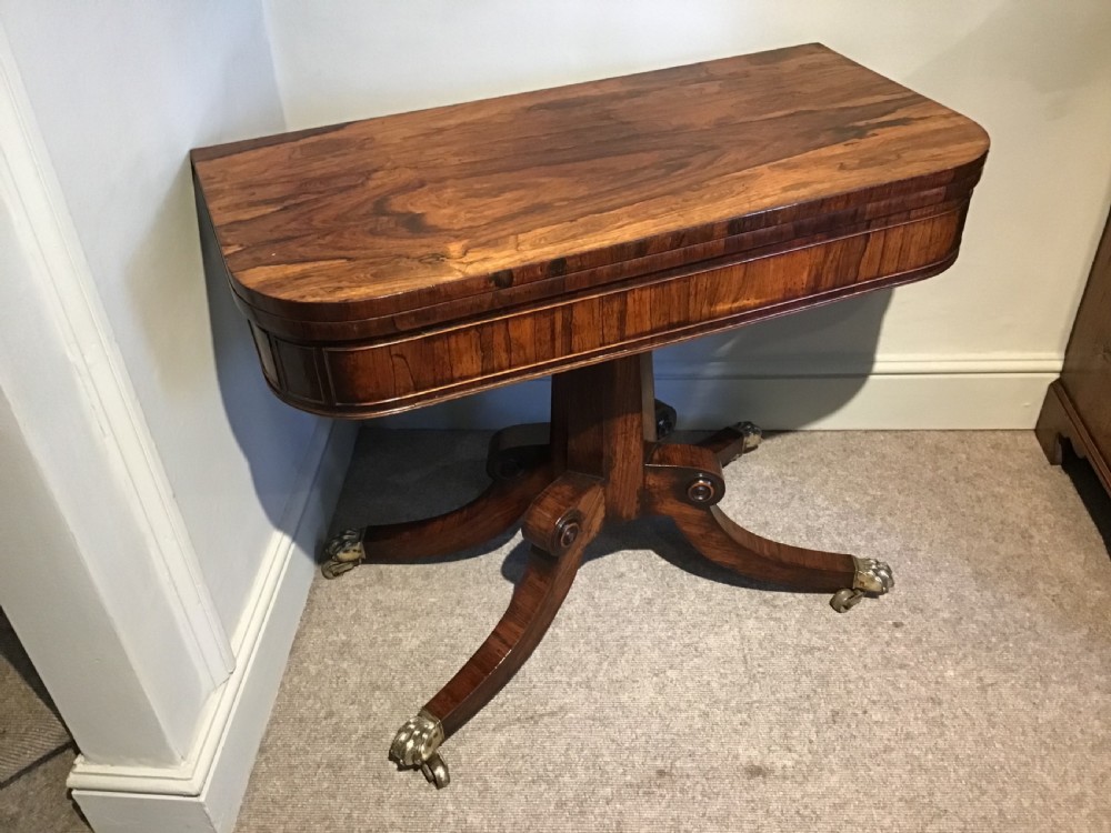 A Regency rosewood card table featuring a rectangular swivel top with rounded corners, supported by a central pedestal column and splayed legs ending in brass lions paw castors.