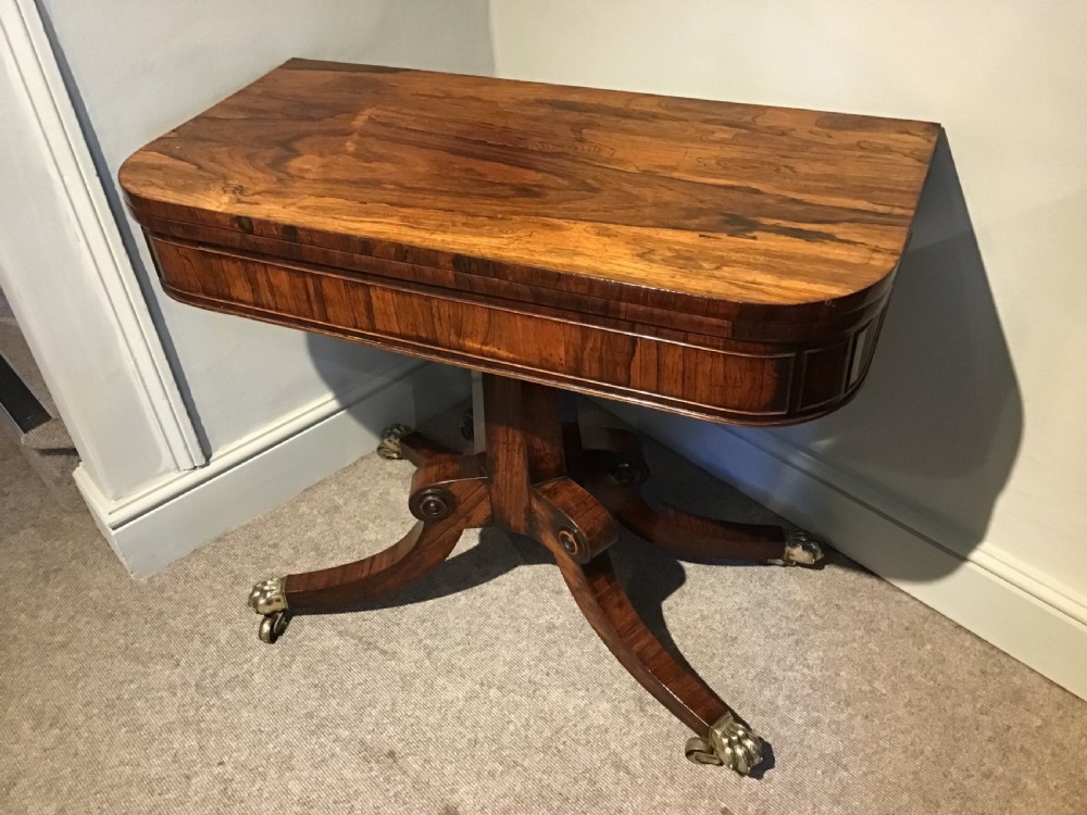 A Regency rosewood card table featuring a rectangular swivel top with rounded corners, supported by a central pedestal column and splayed legs ending in brass lions paw castors. View II