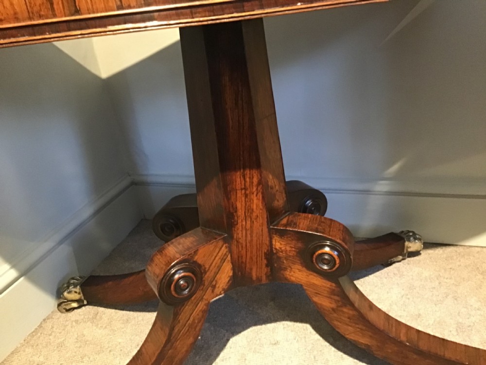 A Regency rosewood card table featuring a rectangular swivel top with rounded corners, supported by a central pedestal column and splayed legs ending in brass lions paw castors. View V