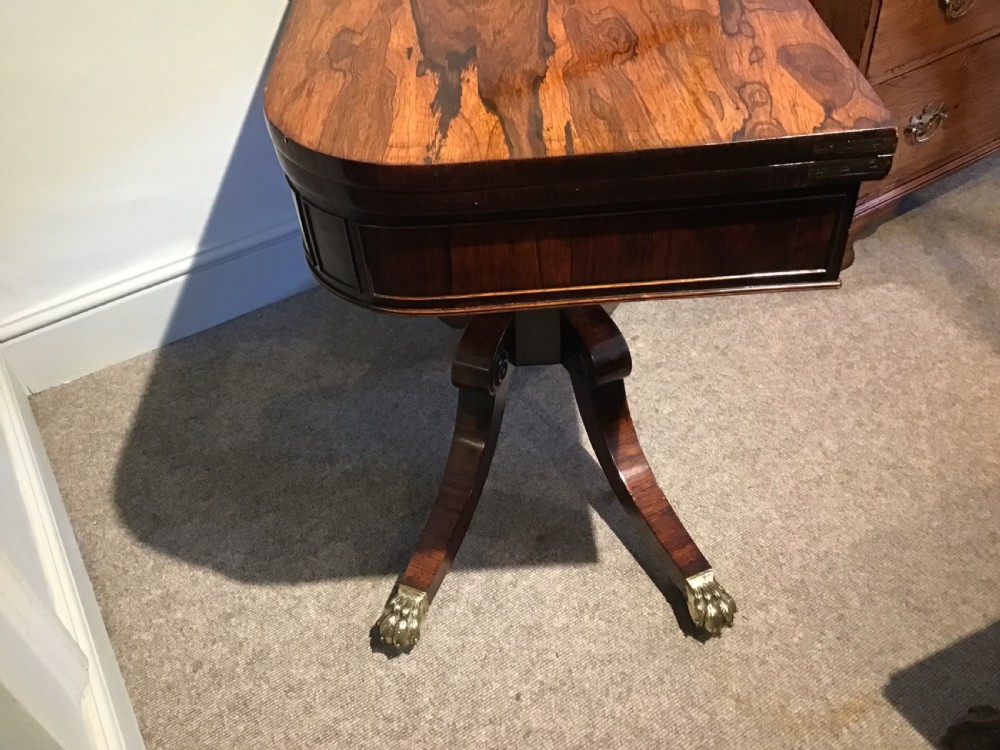 A Regency rosewood card table featuring a rectangular swivel top with rounded corners, supported by a central pedestal column and splayed legs ending in brass lions paw castors. View XI