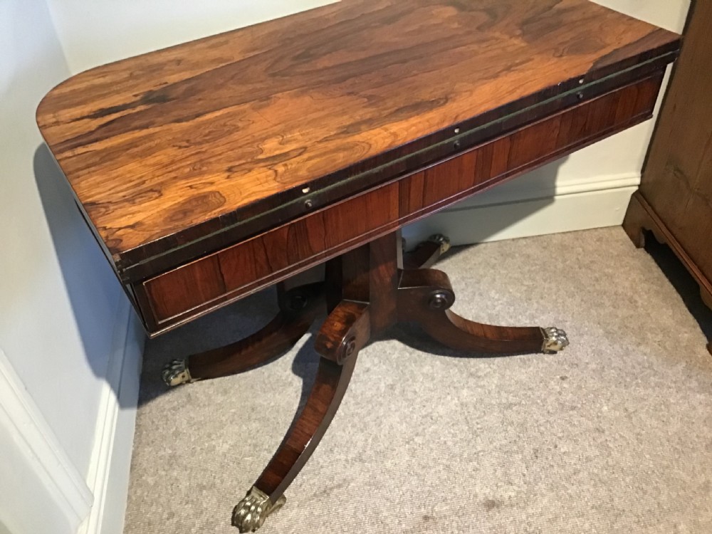 A Regency rosewood card table featuring a rectangular swivel top with rounded corners, supported by a central pedestal column and splayed legs ending in brass lions paw castors. View XII