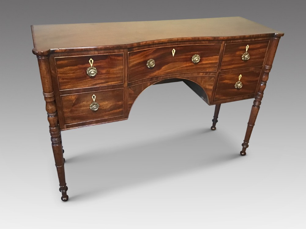 An antique Regency flame mahogany hall table featuring five drawers, brass rosette knobs, kite escutcheons, a central kneehole arch, and slim turned legs.