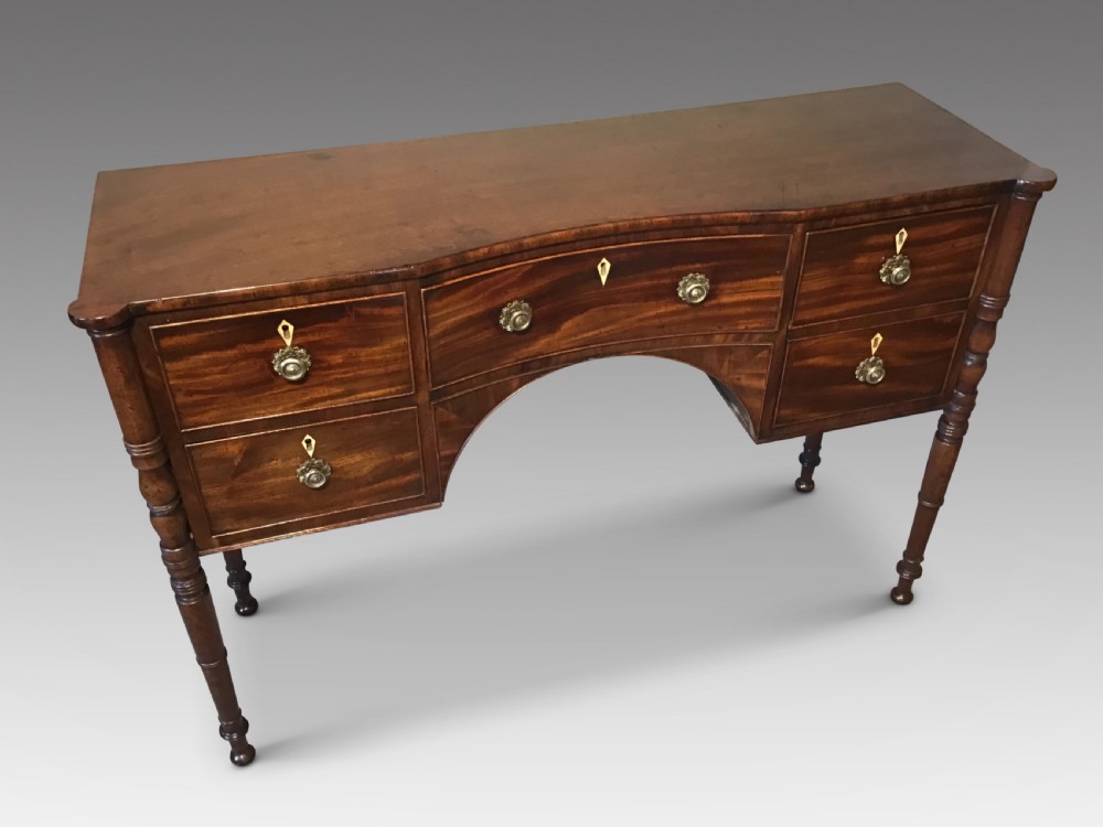 An antique Regency flame mahogany hall table featuring five drawers, brass rosette knobs, kite escutcheons, a central kneehole arch, and slim turned legs. View II