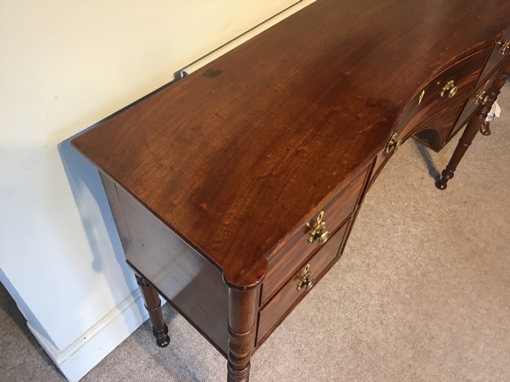 An antique Regency flame mahogany hall table featuring five drawers, brass rosette knobs, kite escutcheons, a central kneehole arch, and slim turned legs. View III