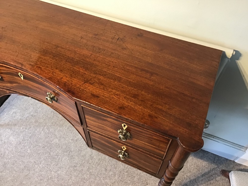 An antique Regency flame mahogany hall table featuring five drawers, brass rosette knobs, kite escutcheons, a central kneehole arch, and slim turned legs. View V
