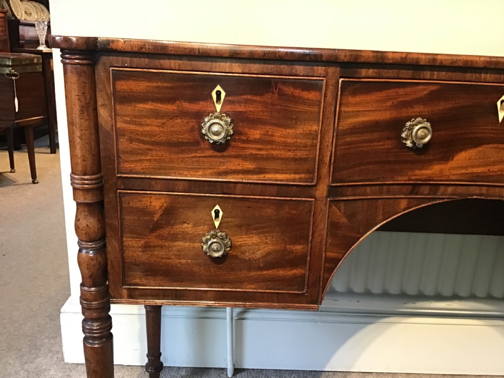 An antique Regency flame mahogany hall table featuring five drawers, brass rosette knobs, kite escutcheons, a central kneehole arch, and slim turned legs. View VI