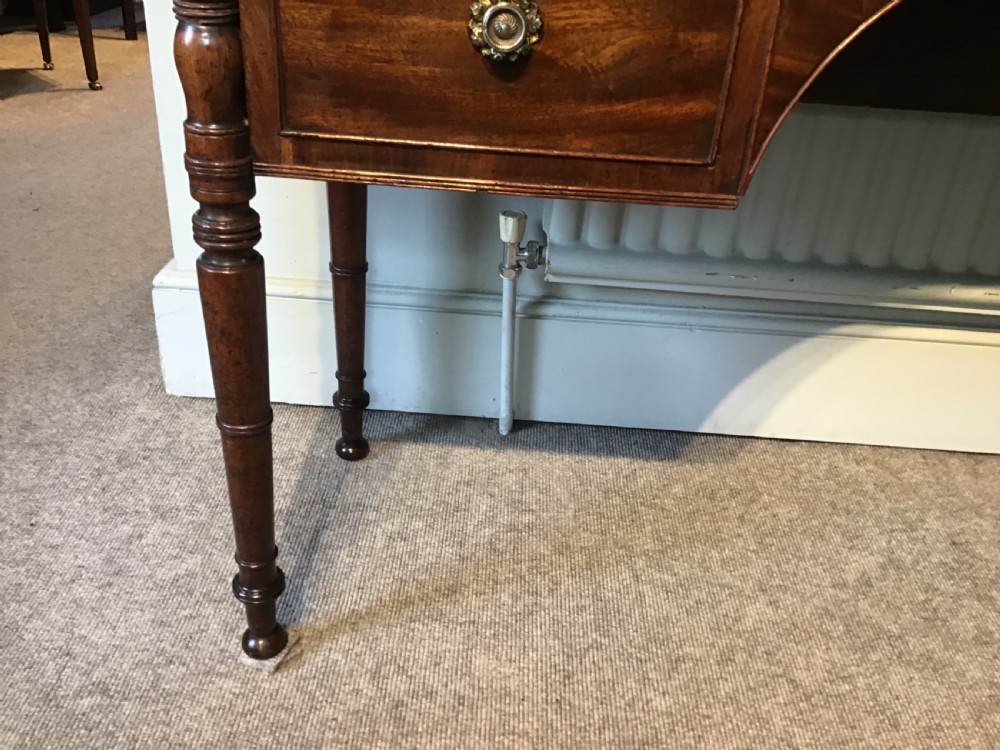 An antique Regency flame mahogany hall table featuring five drawers, brass rosette knobs, kite escutcheons, a central kneehole arch, and slim turned legs. View VII