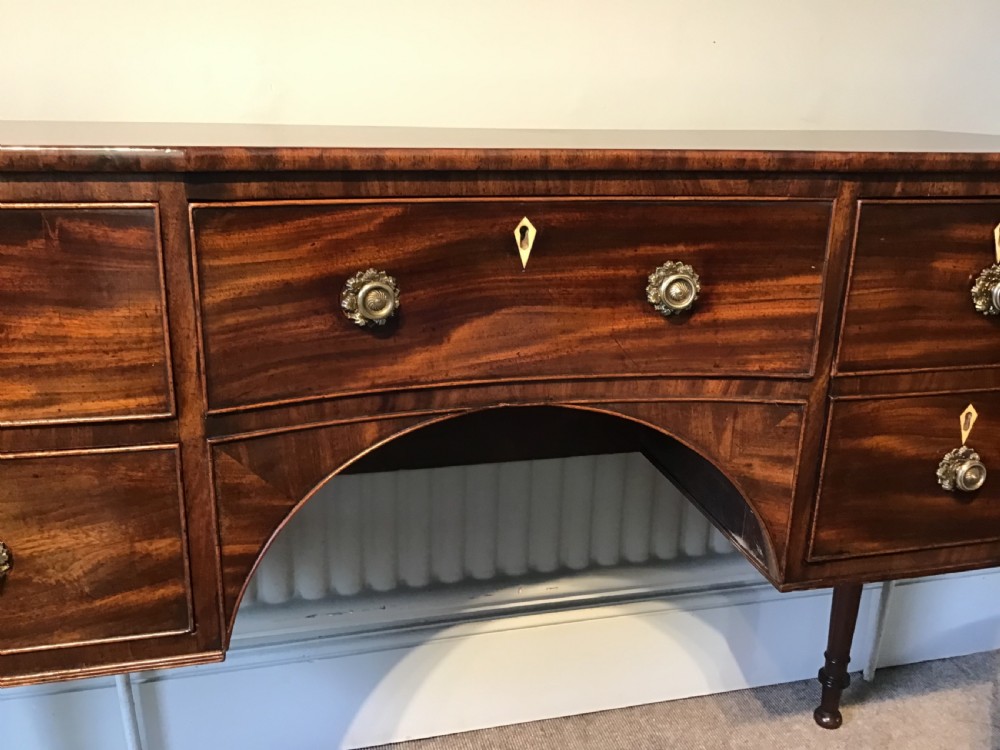 An antique Regency flame mahogany hall table featuring five drawers, brass rosette knobs, kite escutcheons, a central kneehole arch, and slim turned legs. View VIII