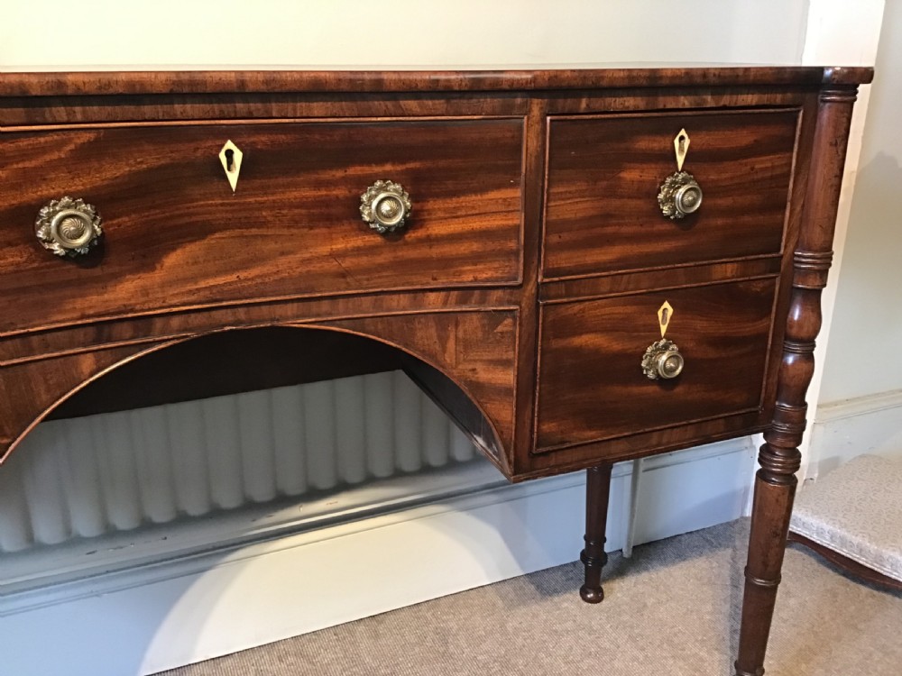 An antique Regency flame mahogany hall table featuring five drawers, brass rosette knobs, kite escutcheons, a central kneehole arch, and slim turned legs. View IX