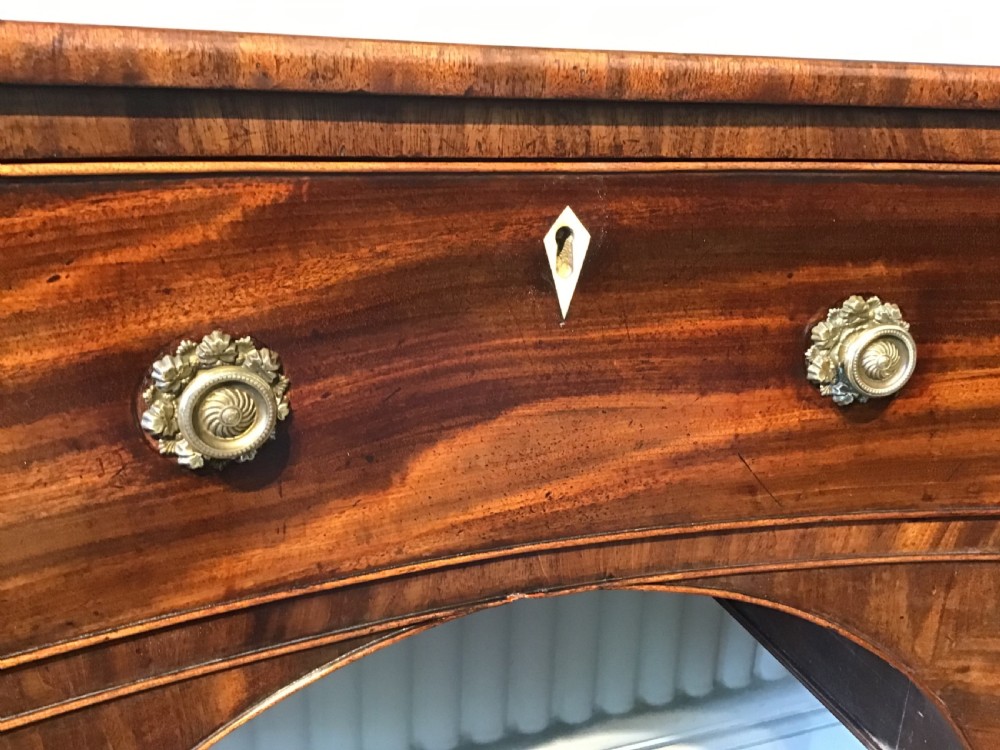 An antique Regency flame mahogany hall table featuring five drawers, brass rosette knobs, kite escutcheons, a central kneehole arch, and slim turned legs. View XI