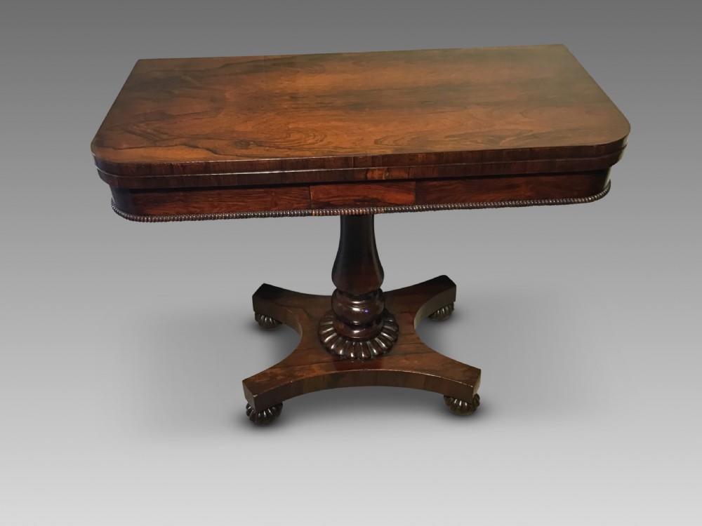 An antique rosewood fold-over card table with a beaded frieze, central turned pedestal stem, and a quadripartite plinth base on carved feet.
