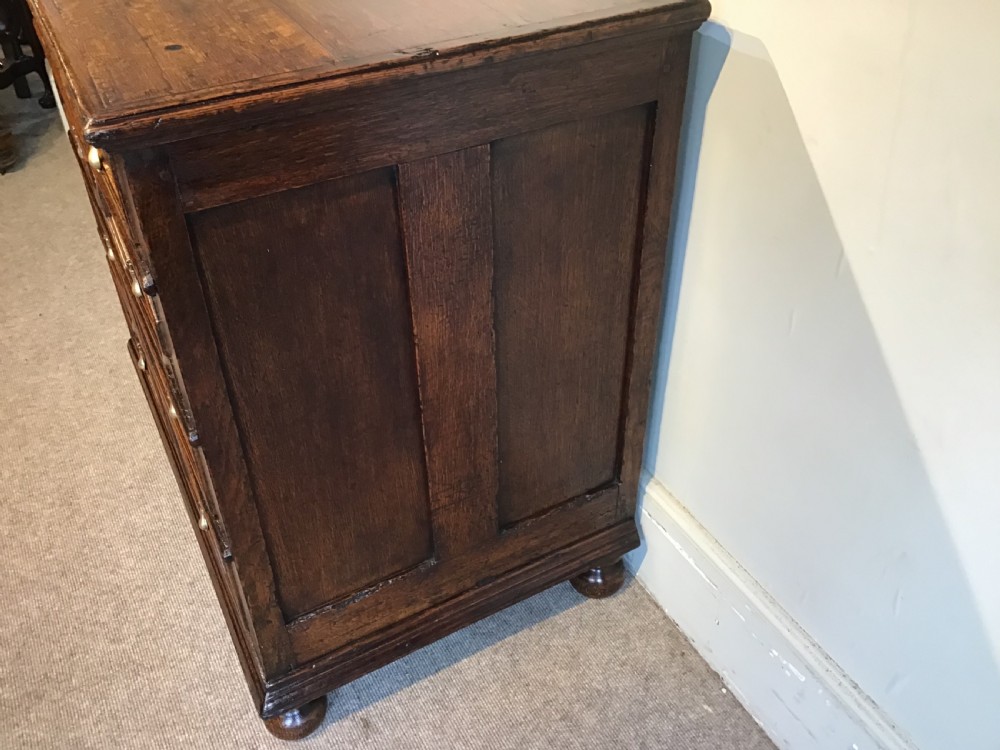An antique dark brown oak chest featuring four drawers with geometric panel mouldings, brass teardrop handles, escutcheons, and bun feet. View V