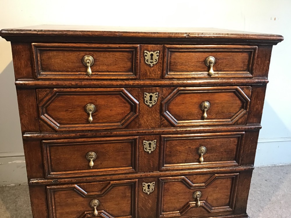 An antique dark brown oak chest featuring four drawers with geometric panel mouldings, brass teardrop handles, escutcheons, and bun feet. View VII