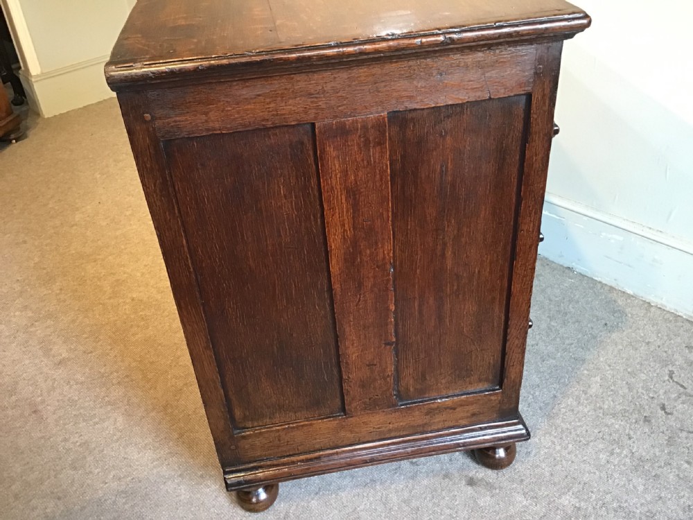 An antique dark brown oak chest featuring four drawers with geometric panel mouldings, brass teardrop handles, escutcheons, and bun feet. View XIII