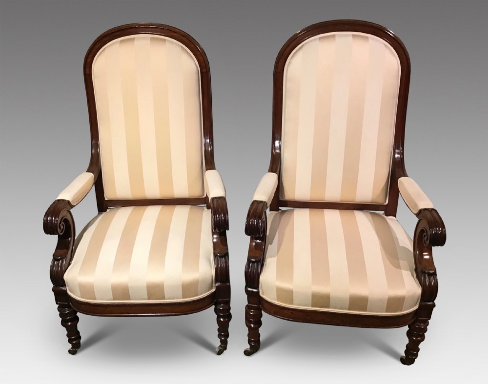 A pair of antique mahogany armchairs featuring tall arched backs, cream striped upholstery, scrolled armrests, and turned legs on castors.