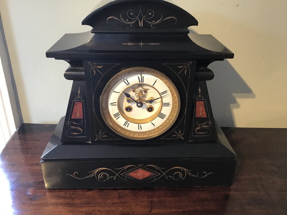 A black antique mantel clock featuring a white circular dial with Roman numerals, an exposed escapement, and gold scrollwork detailing across the case.