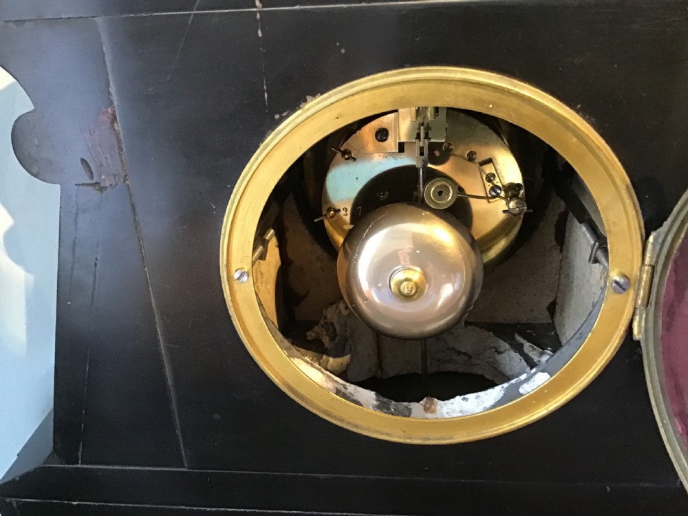 A black antique mantel clock featuring a white circular dial with Roman numerals, an exposed escapement, and gold scrollwork detailing across the case. View IV