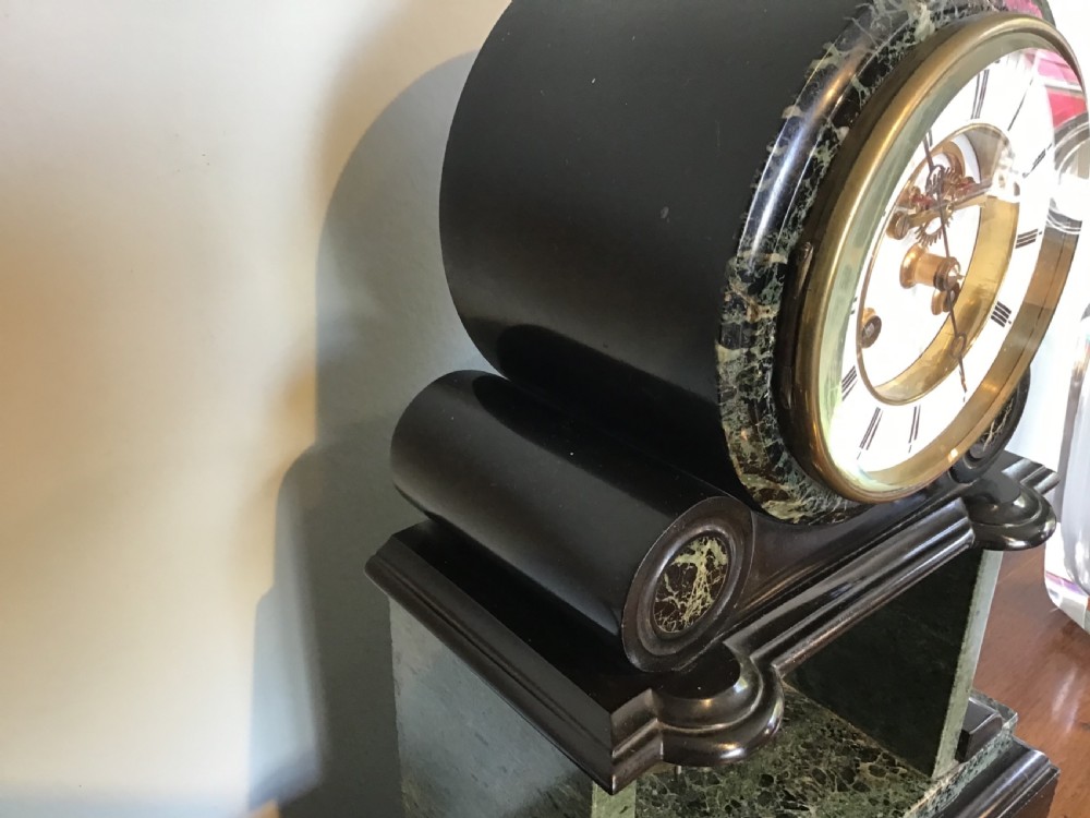 A Victorian black slate and green marble mantel clock featuring a white dial with a visible escapement and a brass pendulum. View V