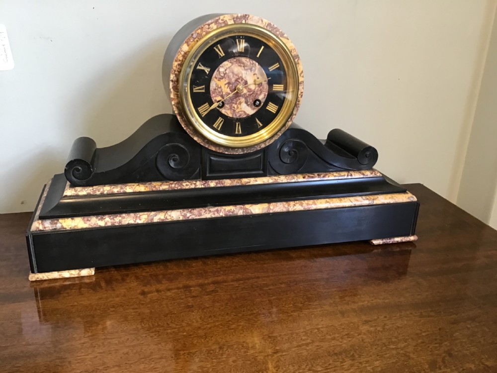 A Victorian black slate and variegated marble drumhead mantel clock featuring a brass-rimmed circular dial set above a scrolled architectural base.