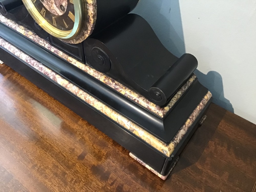 A Victorian black slate and variegated marble drumhead mantel clock featuring a brass-rimmed circular dial set above a scrolled architectural base. View II
