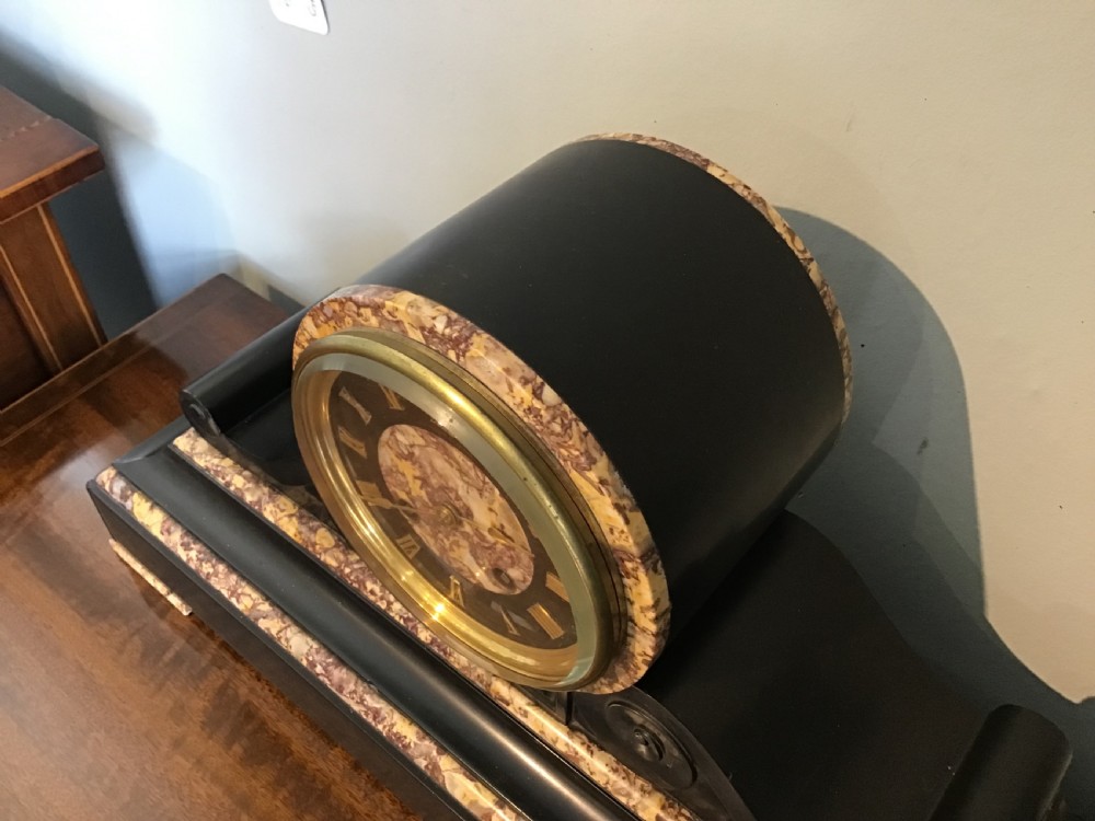 A Victorian black slate and variegated marble drumhead mantel clock featuring a brass-rimmed circular dial set above a scrolled architectural base. View III
