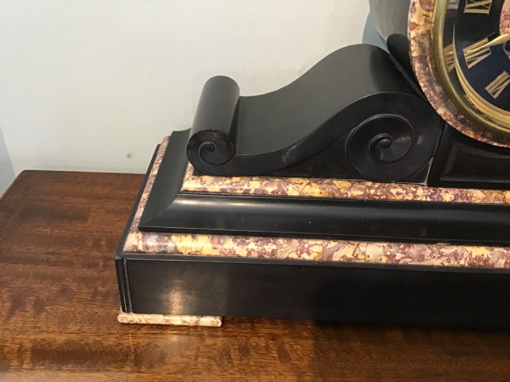 A Victorian black slate and variegated marble drumhead mantel clock featuring a brass-rimmed circular dial set above a scrolled architectural base. View V