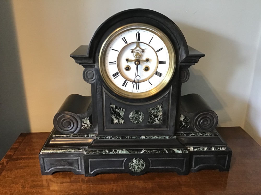 A Victorian black slate mantel clock featuring green marble inlays, scrolled side volutes, and a white enamel dial with an exposed escapement.