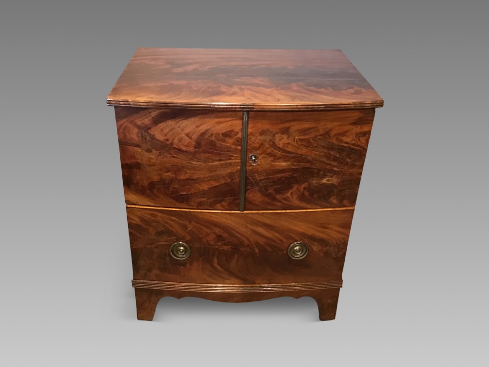An antique bowfront cabinet crafted from flame mahogany veneer with two upper cupboard doors, a lower drawer with round brass ring pulls, and bracket feet.