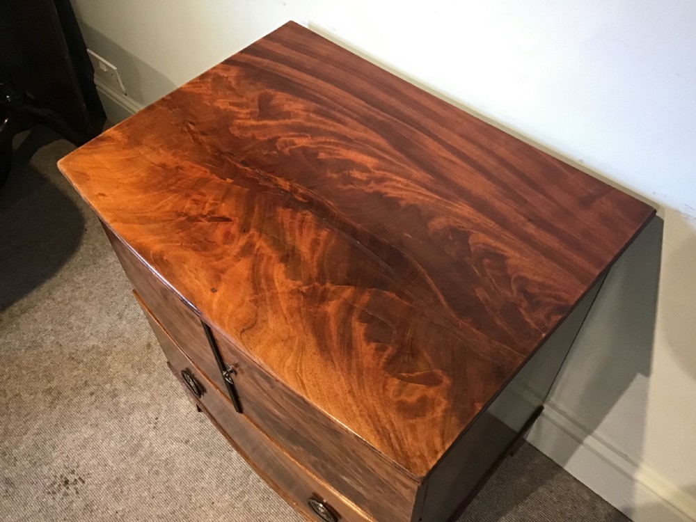 An antique bowfront cabinet crafted from flame mahogany veneer with two upper cupboard doors, a lower drawer with round brass ring pulls, and bracket feet. View III
