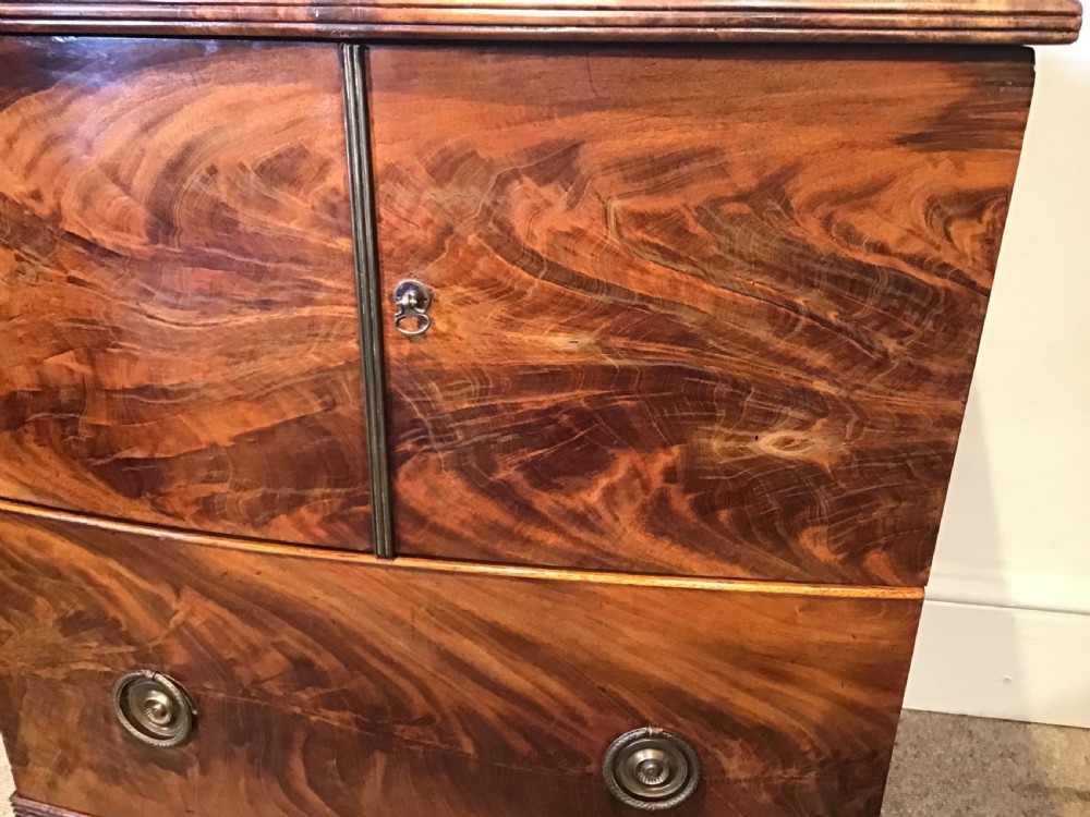 An antique bowfront cabinet crafted from flame mahogany veneer with two upper cupboard doors, a lower drawer with round brass ring pulls, and bracket feet. View V