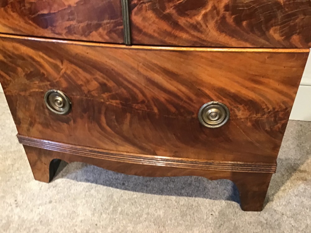 An antique bowfront cabinet crafted from flame mahogany veneer with two upper cupboard doors, a lower drawer with round brass ring pulls, and bracket feet. View VI