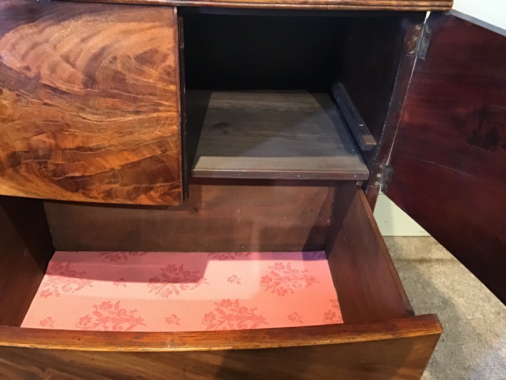 An antique bowfront cabinet crafted from flame mahogany veneer with two upper cupboard doors, a lower drawer with round brass ring pulls, and bracket feet. View VII