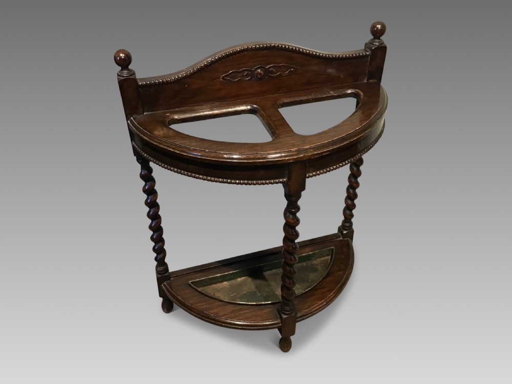 An antique dark oak demilune stick stand featuring three barley twist legs, beaded carved details, two top sections, ball finials, and a fitted metal drip tray.