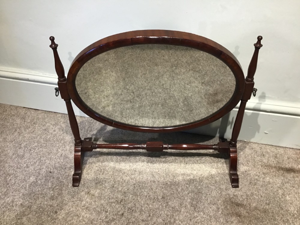 An antique oval mahogany skeleton mirror featuring turned side posts with finials and a swivel mechanism on a low wooden base.
