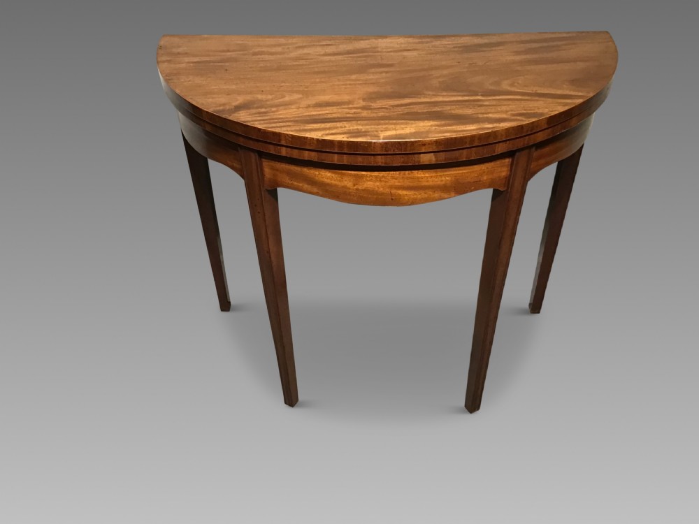 A semi-circular antique mahogany demi-lune card table featuring four square tapered legs and a patterned flame mahogany veneered top surface.