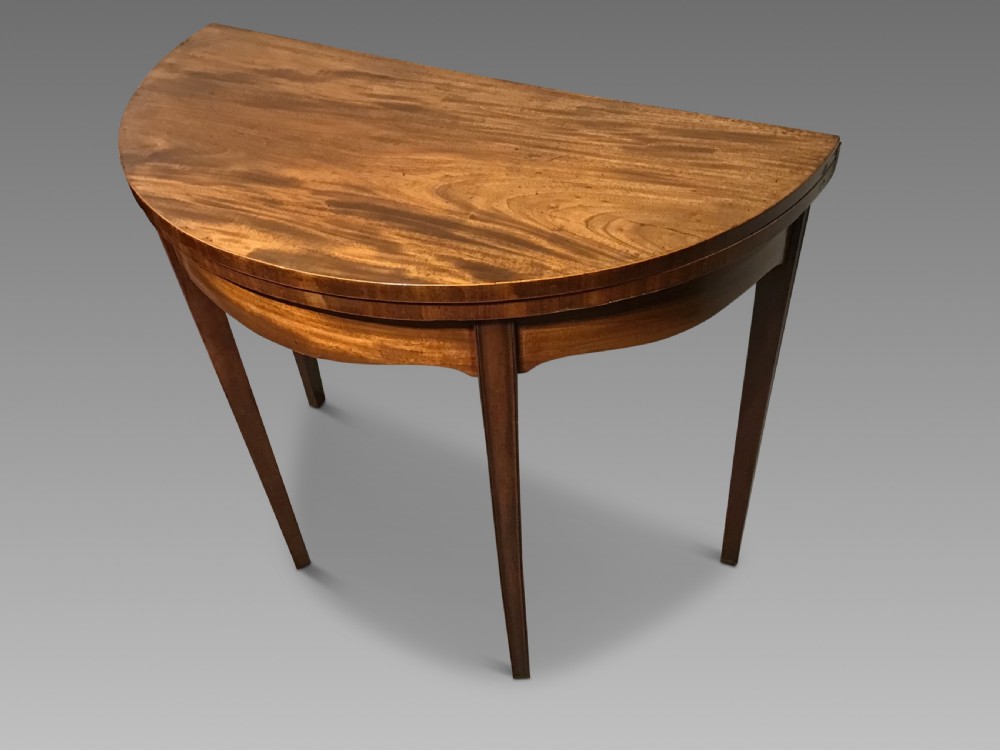 A semi-circular antique mahogany demi-lune card table featuring four square tapered legs and a patterned flame mahogany veneered top surface. View II