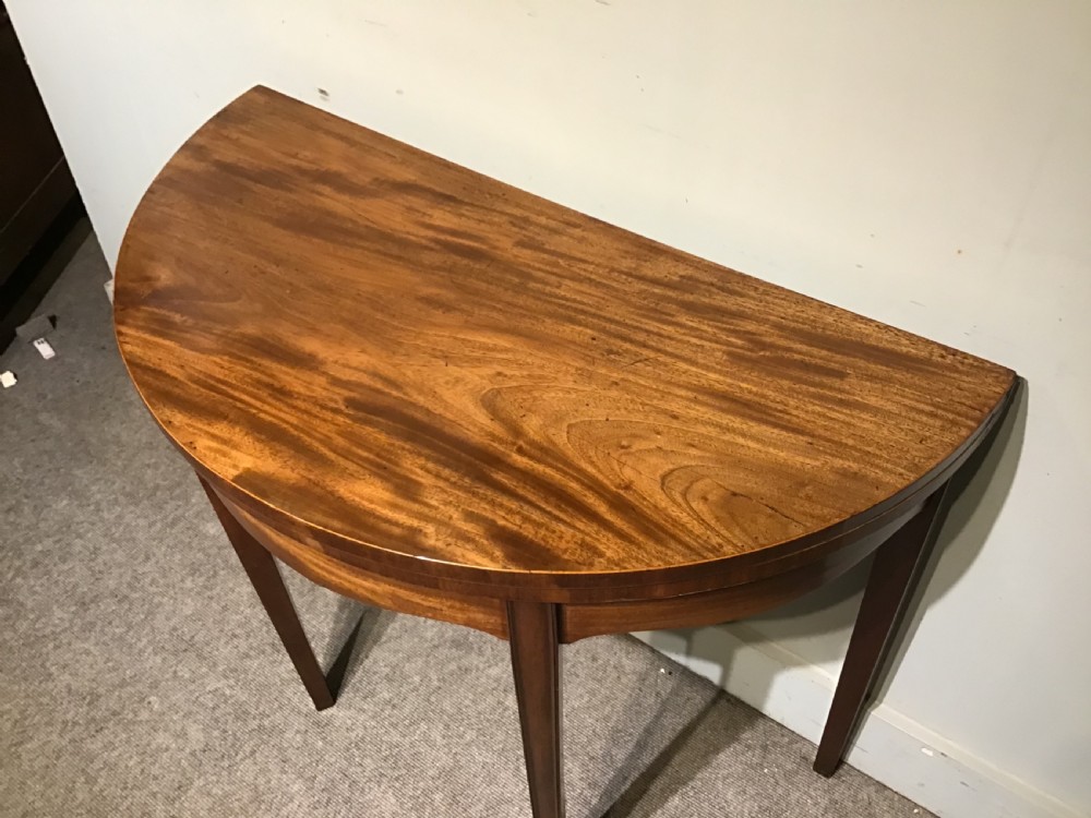 A semi-circular antique mahogany demi-lune card table featuring four square tapered legs and a patterned flame mahogany veneered top surface. View III